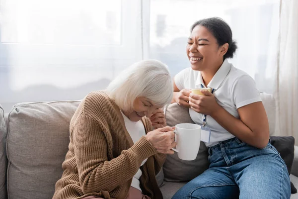 Happy multiracial social worker laughing with senior woman while having tea in living room — Stock Photo