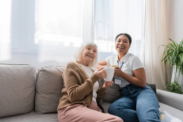 Fröhliche multirassische Sozialarbeiterin sitzt mit einer Seniorin auf dem Sofa und trinkt Tee im Wohnzimmer — Stockfoto