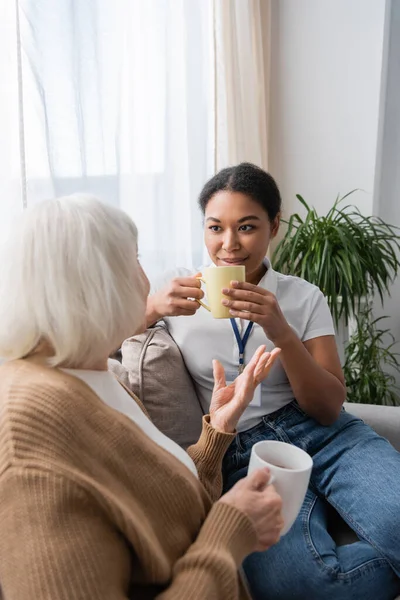 Sonriente trabajador social multirracial charlando con la mujer mayor mientras toma el té en la sala de estar - foto de stock