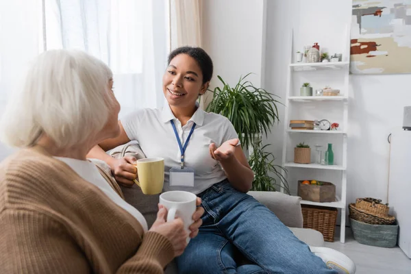 Feliz trabajador social multirracial charlando con una mujer jubilada mientras toma el té en la sala de estar - foto de stock