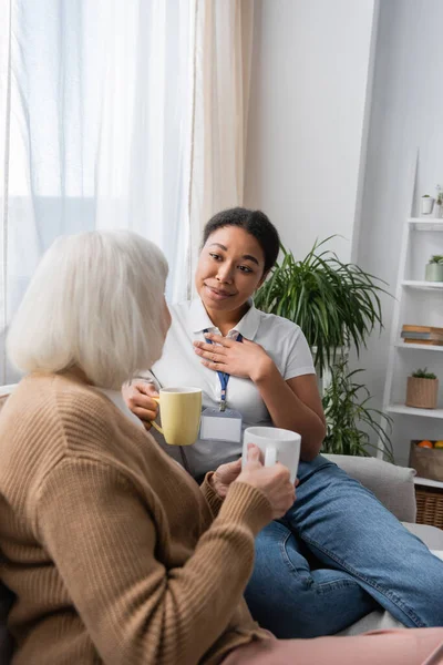 Сентиментальний багаторасовий соціальний працівник балакає з старшою жінкою, маючи чай у вітальні — стокове фото