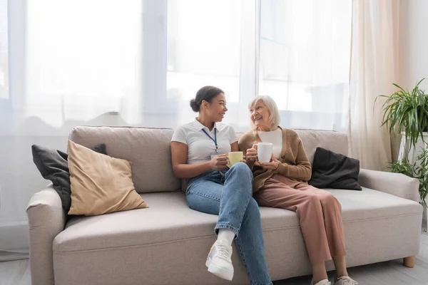 Fröhliche multiethnische Sozialarbeiterin beim Tee mit Seniorin im Wohnzimmer — Stockfoto