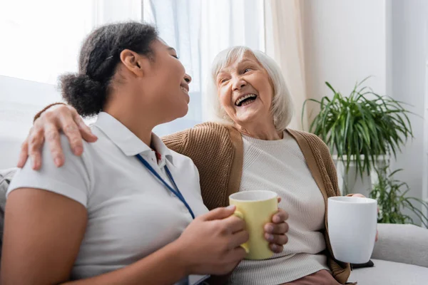 Happy multiracial social worker having tea and laughing with senior woman in living room — Stock Photo