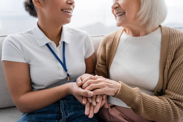 Vista ritagliata di felice assistente sociale multirazziale che si tiene per mano con la donna anziana in soggiorno — Foto stock