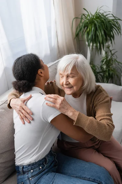 Trabajador social multirracial abrazándose con una mujer mayor mientras está sentado en el sofá en la sala de estar - foto de stock