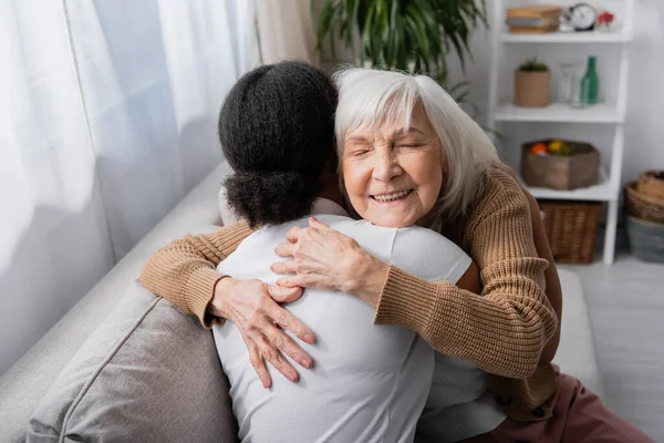 Счастливая пожилая женщина с закрытыми глазами обнимает кудрявого социального работника в гостиной — стоковое фото
