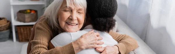 Mujer mayor feliz con los ojos cerrados abrazando trabajador social multirracial en la sala de estar, pancarta - foto de stock