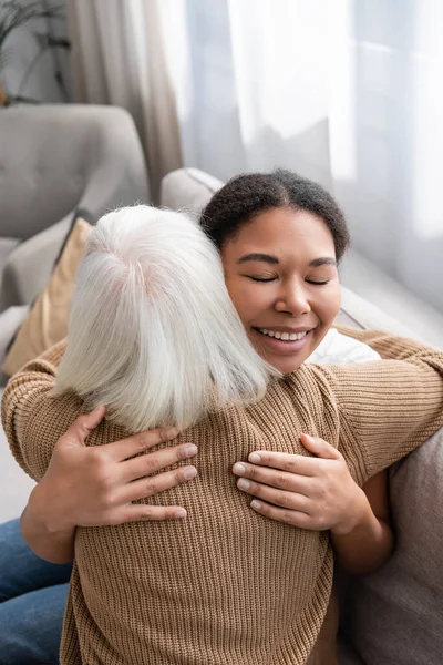 Щасливий багаторасовий соціальний працівник з закритими очима обіймається з старшою жінкою у вітальні — стокове фото