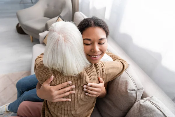 Cheerful multiracial social worker with closed eyes hugging with senior woman in living room — Stock Photo
