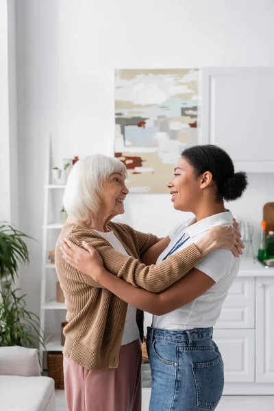 Вид збоку щасливого багаторасового соціального працівника обіймається з старшою жінкою у вітальні — стокове фото