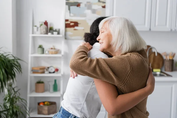 Cheerful senior woman with closed eyes hugging multiracial social worker — Stock Photo