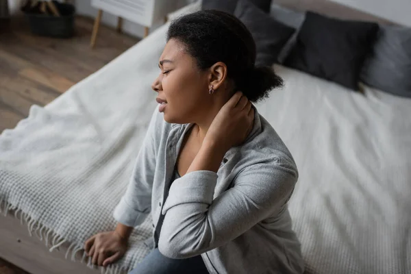 Side view of upset multiracial woman with depression touching neck while sitting on bed — Stock Photo