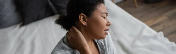 Vue latérale de triste femme multiraciale touchant le cou près du lit à la maison, bannière — Photo de stock