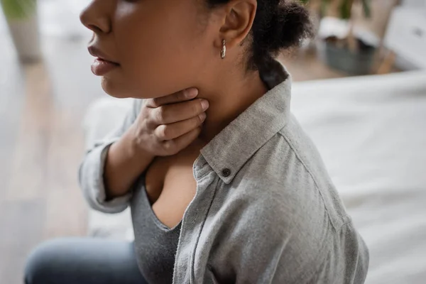 Cropped view of young multiracial woman touching throat while sitting on blurred bed at home — Stock Photo