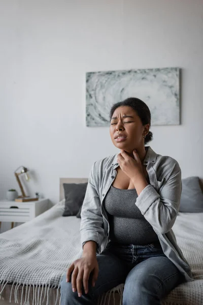 Gestresste multiethnische Frau berührt Halsschmerzen, während sie zu Hause auf dem Bett sitzt — Stockfoto