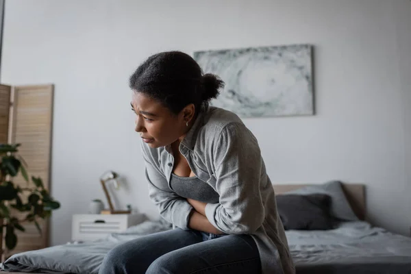 Tired multiracial woman feeling cramp in stomach while sitting on bed — Stock Photo