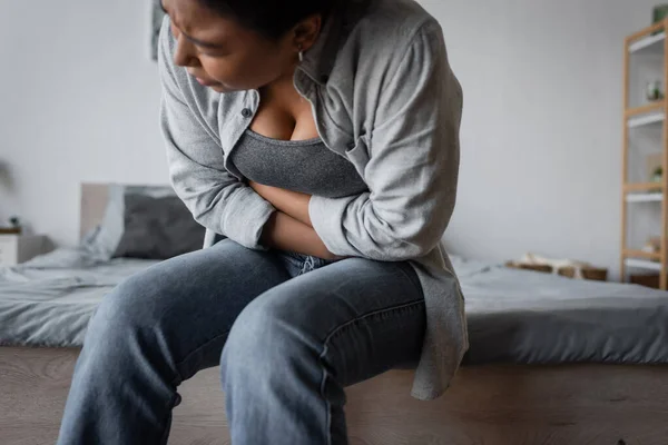 Displeased multiracial woman touching aching stomach on bed at home — Stock Photo