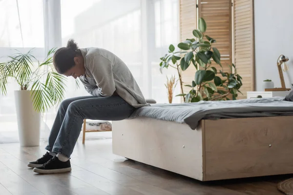 Brünette multirassische Frau leidet unter Schmerzen, während sie im Schlafzimmer auf dem Bett sitzt — Stockfoto
