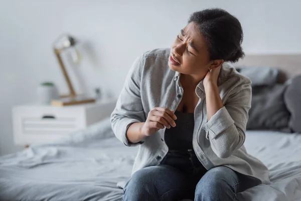 Tired multiracial woman touching neck and suffering from pain in bedroom at home — Stock Photo