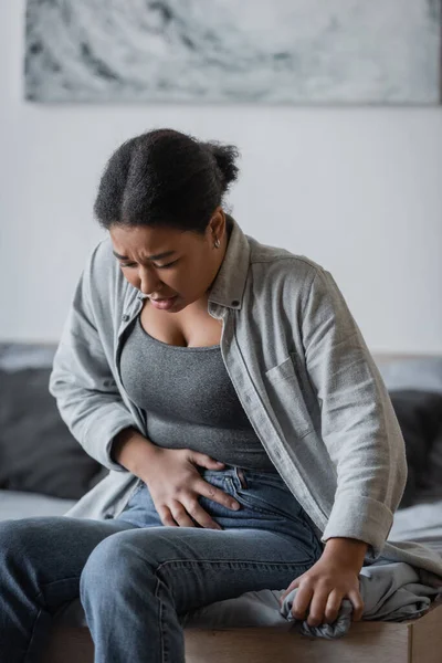 Verspannte multirassische Frau berührt Bauch, während sie zu Hause auf dem Bett sitzt — Stockfoto