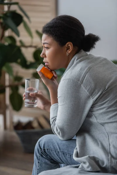 Vista laterale di donna multirazziale triste che tiene pillole e acqua mentre si siede sul letto a casa — Foto stock