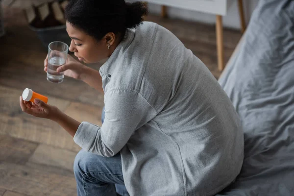 Blick von oben auf traurige multirassische Frau, die Pillen betrachtet und ein Glas Wasser im Schlafzimmer hält — Stockfoto