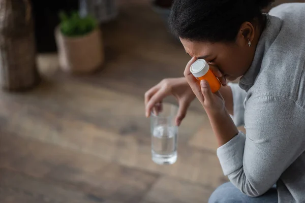 Незадоволена мультирасова жінка тримає таблетки і розмиту склянку води вдома — стокове фото