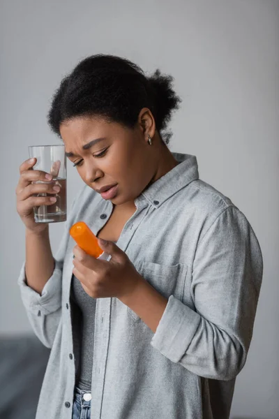 Donna multirazziale sconvolta con pillole e bicchiere d'acqua a casa — Foto stock
