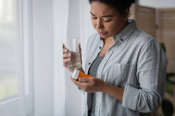 Disappointed multiracial woman with depression holding pills and water near curtain at home — Stock Photo