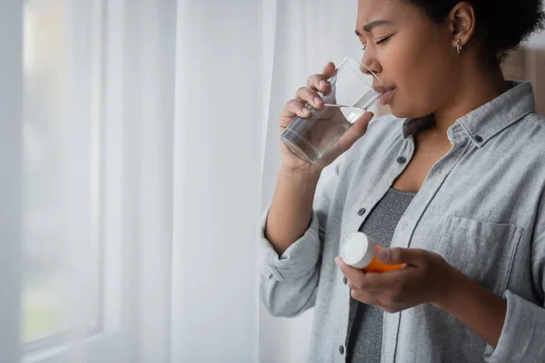 Multiracial woman with depression taking pill and drinking water at home — Stock Photo