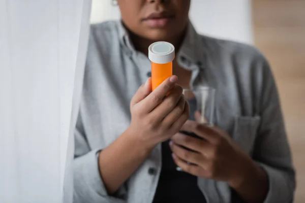 Vista cortada de mulher multirracial desfocada segurando pílulas antidepressivas e água perto de cortina em casa — Fotografia de Stock