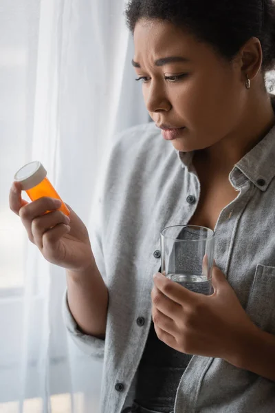 Mujer joven multirracial con depresión sosteniendo pastillas y vaso de agua en casa - foto de stock