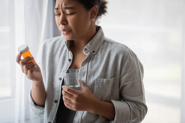Disappointed multiracial woman with closed eyes holding pills and water at home — Stock Photo
