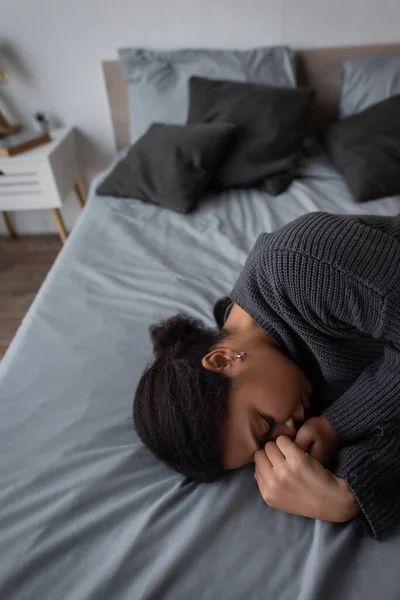 Depressed multiracial woman in knitted sweater lying on bed in blurred bedroom — Stock Photo