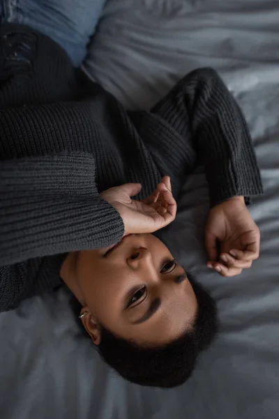 Visão superior da mulher multirracial descontente em suéter deitado na cama em casa — Fotografia de Stock