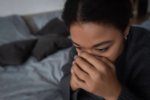 Мультирасовая женщина с психическими проблемами, закрывающая лицо, сидя дома на кровати — стоковое фото