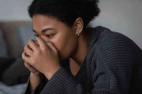 Sad multiracial woman in knitted sweater looking away in bedroom — Stock Photo