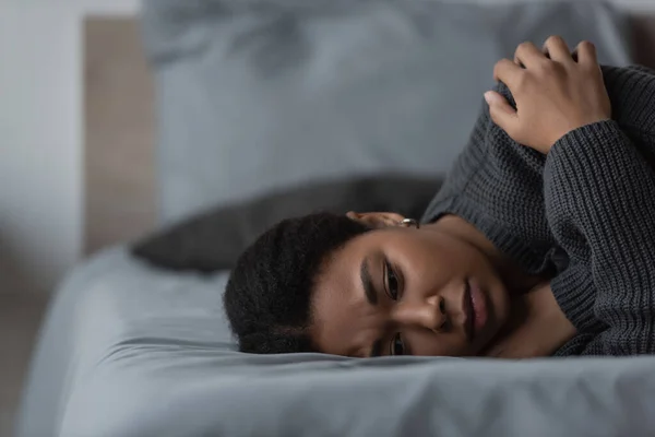 Frustrated multiracial woman in sweater lying on bed at home — Stock Photo