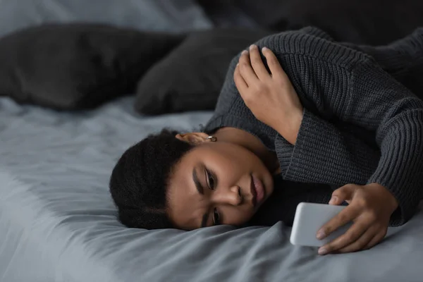 Multiracial woman with depression using smartphone while lying on bed at home — Stock Photo