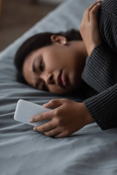 Blurred multiracial woman with depression using smartphone while lying on bed at home — Stock Photo