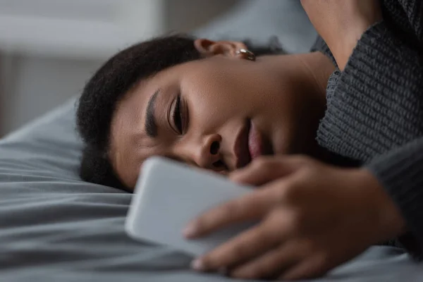 Frustrated multiracial woman using blurred cellphone while lying on bed at home — Stock Photo