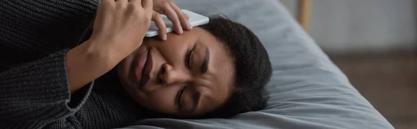 Aufgebrachte multiethnische Frau spricht auf Smartphone, während sie zu Hause im Bett liegt, Banner — Stockfoto
