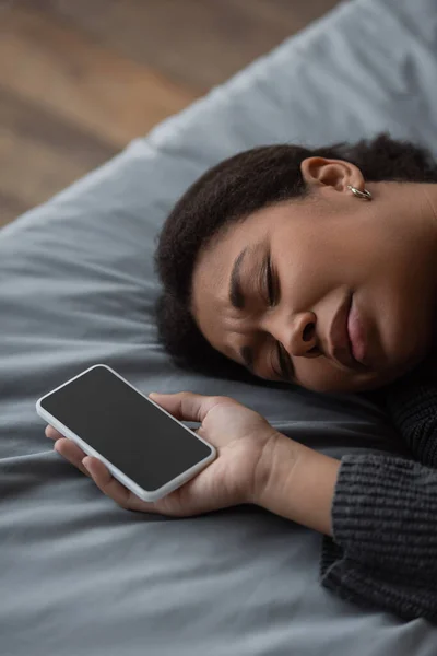 Mujer deprimida multirracial sosteniendo teléfono inteligente con pantalla en blanco mientras está acostado en la cama - foto de stock
