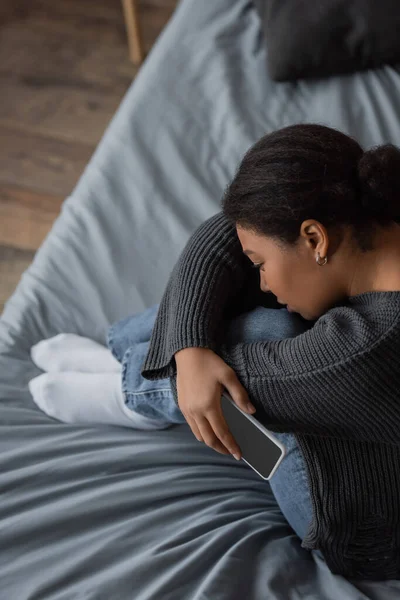 Vue grand angle d'une femme multiraciale frustrée tenant un téléphone portable alors qu'elle était assise sur son lit à la maison — Photo de stock