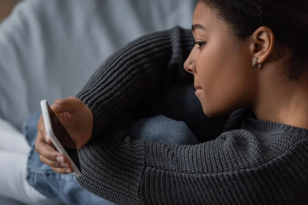 Vista lateral de la deprimida mujer multirracial sosteniendo teléfono inteligente con pantalla en blanco en el dormitorio - foto de stock