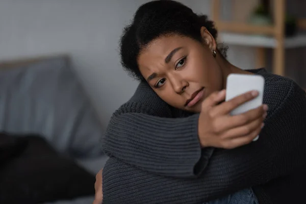 Heartbroken multiracial woman in sweater using blurred smartphone in blurred bedroom — Stock Photo