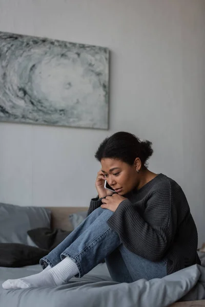 Frustrated multiracial woman calling in helpline while sitting on bed at home — Stock Photo