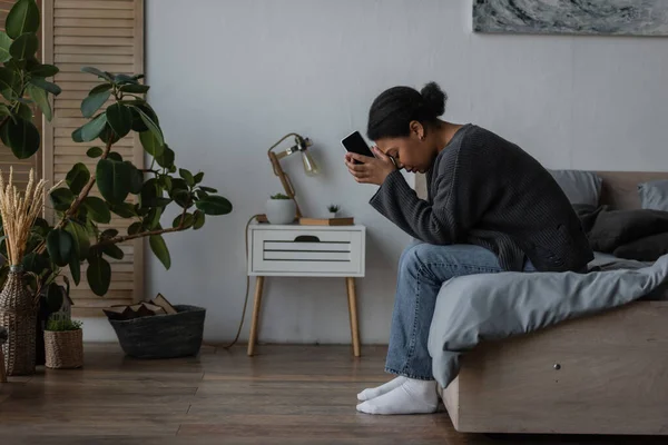 Vue latérale de la femme multiraciale triste tenant smartphone tout en étant assis sur le lit à la maison — Photo de stock