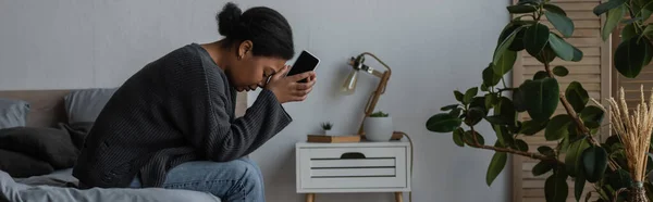 Vista lateral de la deprimida mujer multirracial sosteniendo el teléfono móvil mientras está sentada en la cama en casa, pancarta - foto de stock