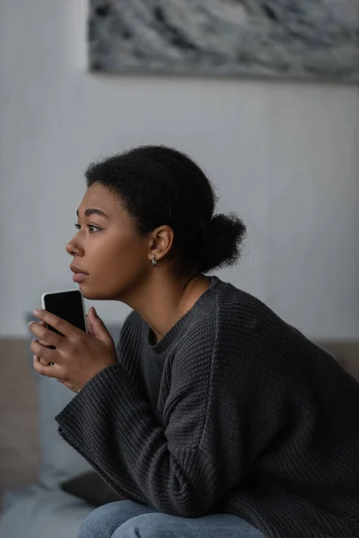 Femme multiraciale au cœur brisé avec dépression tenant téléphone portable dans la chambre — Photo de stock